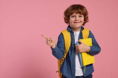 Sırt çantalı ve kitaplı mutlu okul çocuğu pembe arka plandaki bir şeyi işaret ediyor, mesaj için yer var.