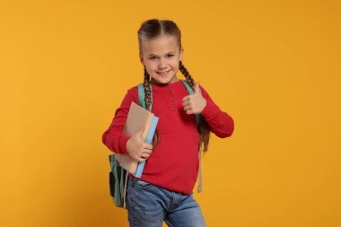 Sırt çantalı ve kitaplı mutlu okul kızı turuncu arka planda başparmak hareketi gösteriyor.
