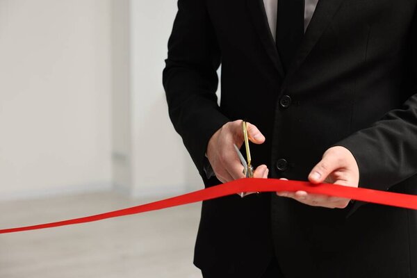 Man cutting red ribbon with scissors indoors, closeup. Space for text