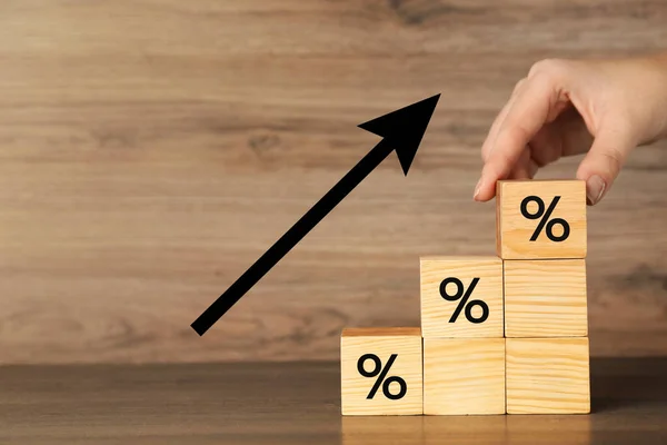 Mortgage rate rising illustrated by upward arrow. Woman putting cube with percent sign on other ones at wooden table, closeup
