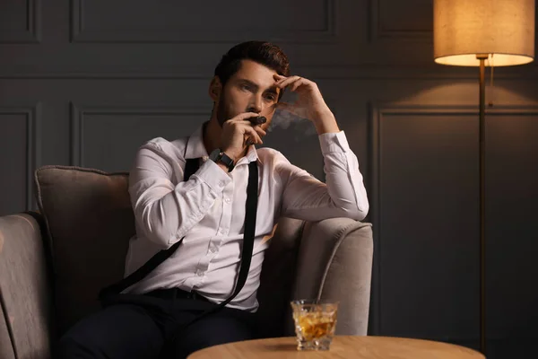 Tired man smoking cigar and resting at home in evening. Glass of whiskey on table near him