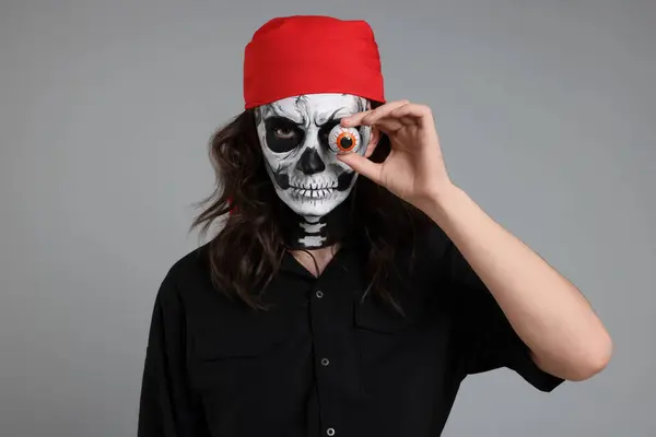 Man in scary pirate costume with skull makeup and decorative eyeball on light grey background. Halloween celebration