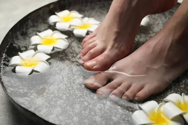 Woman soaking her feet in bowl with water and flowers on floor, closeup. Spa treatment