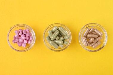 Sarı arka planda cam kaselerde farklı vitamin hapları var.