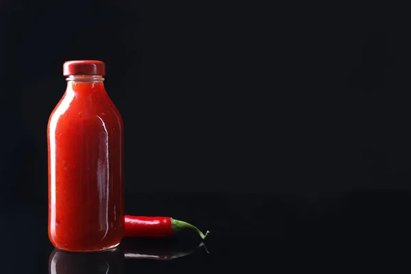 Spicy chili sauce in bottle and pepper against dark background, space for text
