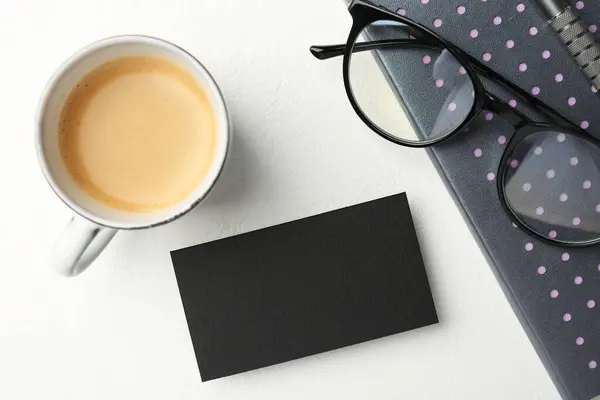 Blank black business card, cup of coffee, glasses and stationery on white table, top view. Mockup for design