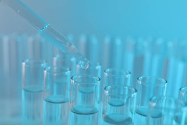 Laboratuvar analizi. Açık mavi arkaplanda pipetten cam test tüpüne sıvı damlatılıyor.