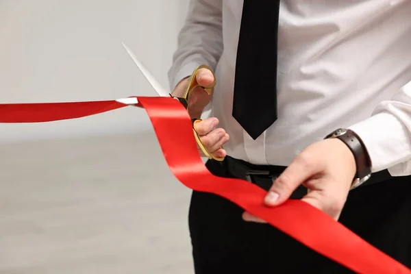 Man cutting red ribbon with scissors on blurred background, closeup