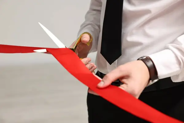 Man cutting red ribbon with scissors on blurred background, closeup