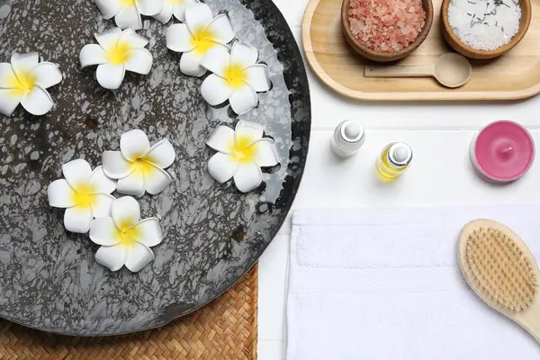 Çiçekli bir kase su ve beyaz ahşap masada farklı spa malzemeleri.