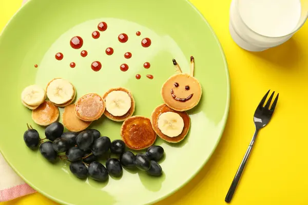 Creative serving for kids. Plate with cute caterpillar made of pancakes, grapes and banana on yellow background, flat lay