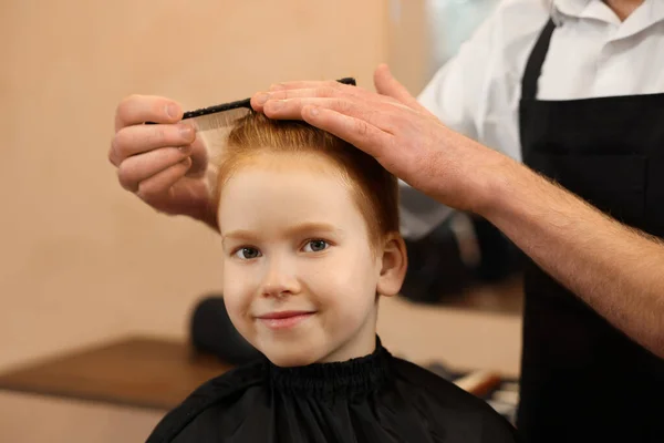 Professional hairdresser combing boy\'s hair in beauty salon, closeup