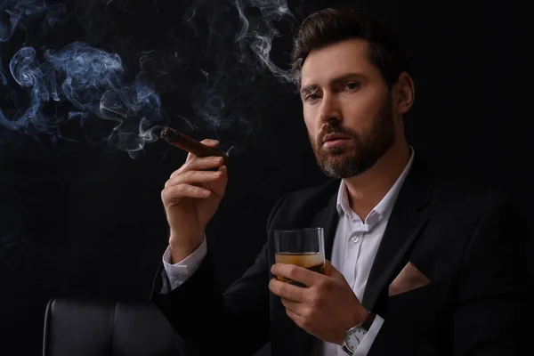 Handsome man in elegant suit with glass of whiskey smoking cigar on black background
