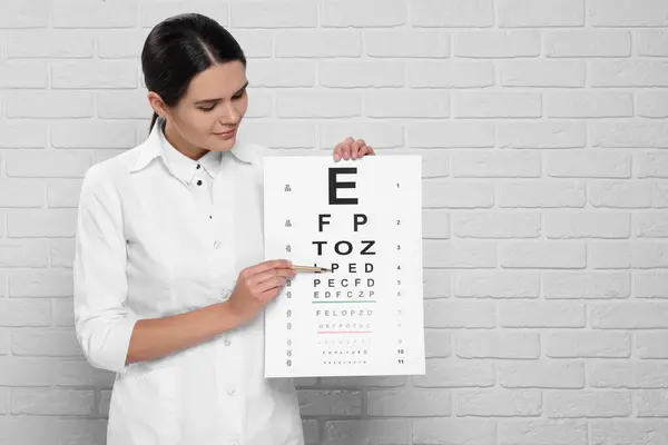 Ophthalmologist pointing at vision test chart near white brick wall, space for text