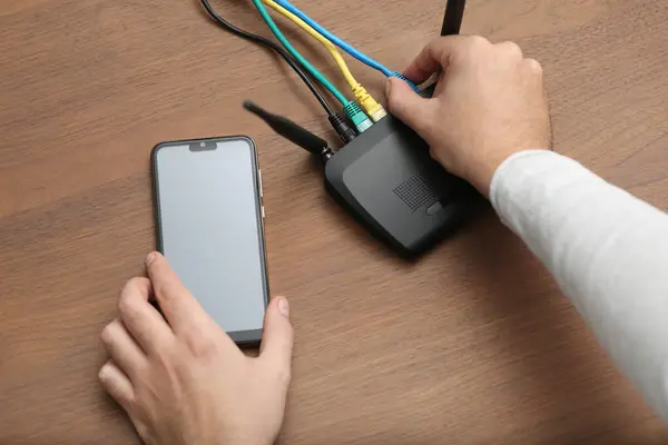 Man with smartphone connecting to internet via Wi-Fi router at wooden table, closeup