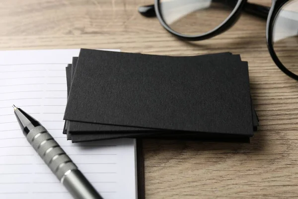 Blank black business cards, notebook, glasses and pen on wooden table, closeup. Mockup for design