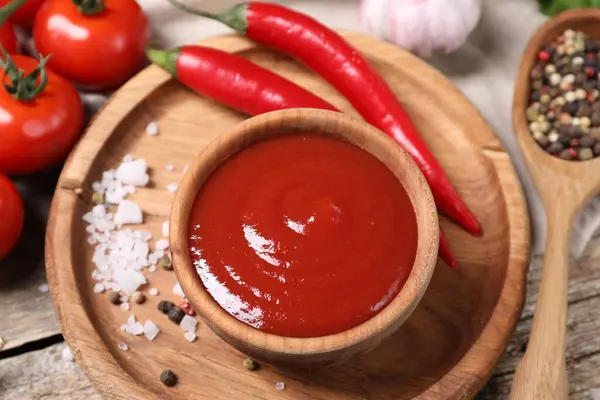 Delicious ketchup in bowl, chili pepper and spices on wooden table, closeup. Tomato sauce