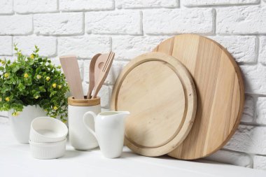 Kesme tahtaları, mutfak aletleri, bulaşık takımları ve ev bitkileri tuğla duvarın yanındaki beyaz masada.