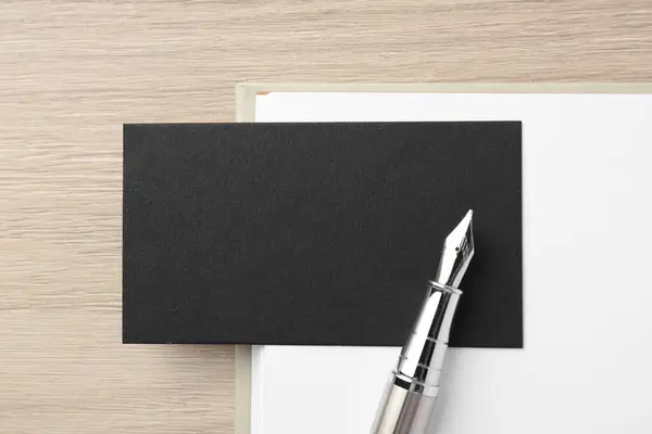 Blank black business card, fountain pen and notebook on wooden table, top view. Mockup for design