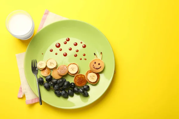 Creative serving for kids. Plate with cute caterpillar made of pancakes, grapes and banana on yellow background, flat lay. Space for text