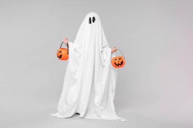 Beyaz hayalet kostümlü çocuk açık gri arka planda balkabağı kovaları tutuyor. Cadılar Bayramı