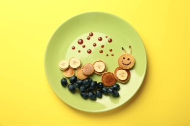 Çocuklar için yaratıcı hizmet. Sarı zemin üzerinde krep, üzüm ve muzdan yapılmış şirin tırtıllı bir tabak.