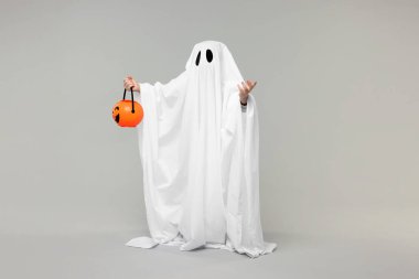 Beyaz hayalet kostümlü çocuk açık gri arka planda balkabağı kovası tutuyor. Cadılar Bayramı