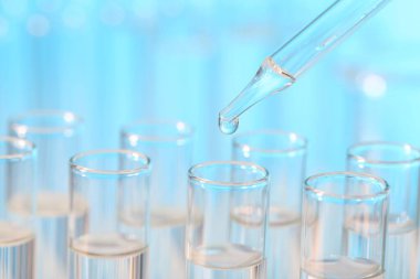 Laboratuvar analizi. Açık mavi arkaplanda pipetten cam test tüpüne sıvı damlatılıyor.