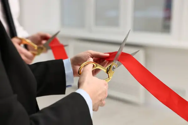 People cutting red ribbon with scissors indoors, closeup