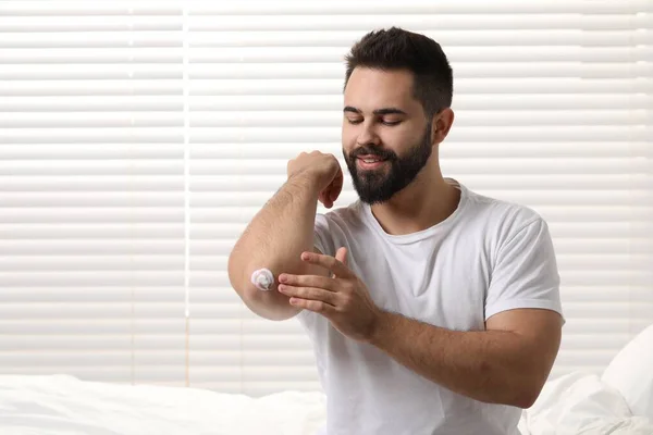 Man with dry skin applying cream onto his elbow indoors, space for text