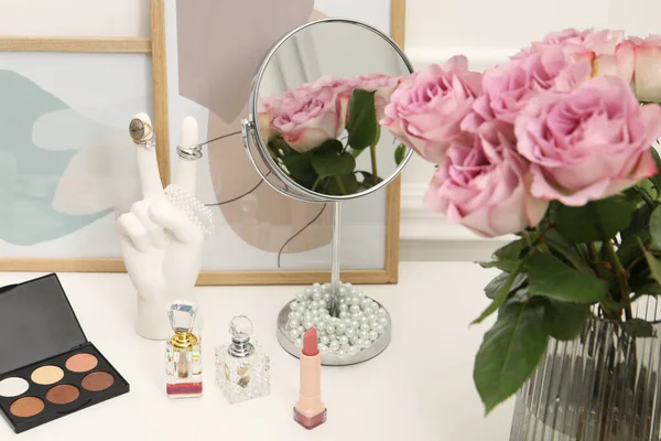 Mirror, cosmetic products, perfumes and vase with pink roses on white dressing table