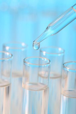 Laboratuvar analizi. Açık mavi arkaplanda pipetten cam test tüpüne sıvı damlatılıyor.
