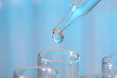 Laboratuvar analizi. Açık mavi arkaplanda pipetten cam test tüpüne sıvı damlatılıyor.