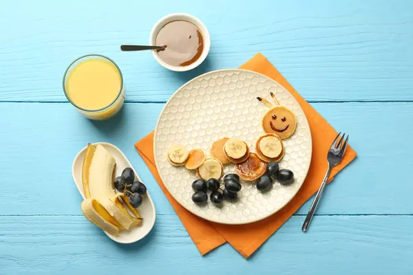 Creative serving for kids. Plate with cute caterpillar made of pancakes, grapes and banana on light blue wooden table, flat lay