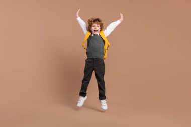 Kahverengi arka planda atlayan sırt çantalı mutlu okul çocuğu.