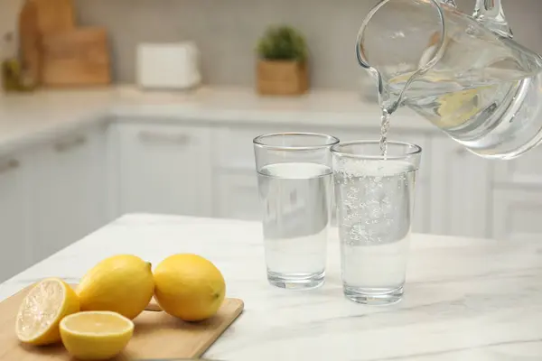 Pouring water from jug into glass on white table in kitchen, closeup