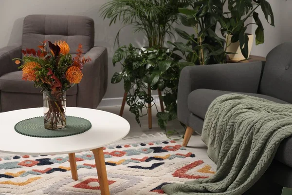 Vase with bouquet of beautiful leucospermum flowers on coffee table, sofa, armchair and houseplants in room