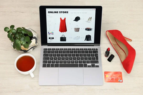 Online store. Laptop with open website, credit card, red high-heeled shoe and lipstick on wooden surface, above view