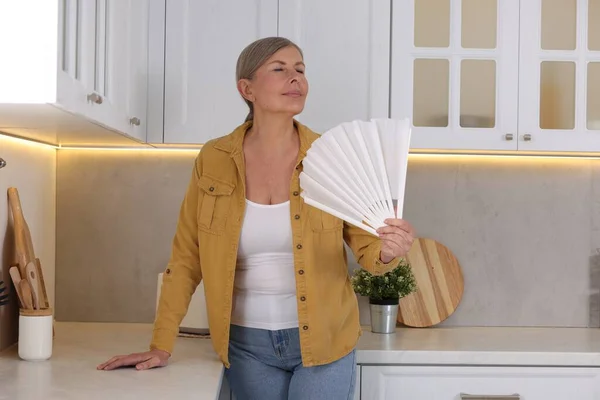 Menopause. Woman waving hand fan to cool herself during hot flash in kitchen