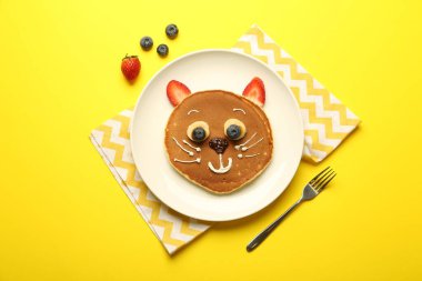 Çocuklar için yaratıcı hizmet. Sarı arka planda krep, böğürtlen, krema, muz ve çikolata ezmesinden yapılmış şirin bir kedi tabağı.