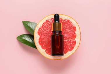 Bir şişe greyfurt yağı, yapraklar ve pembe arka planda taze meyve.