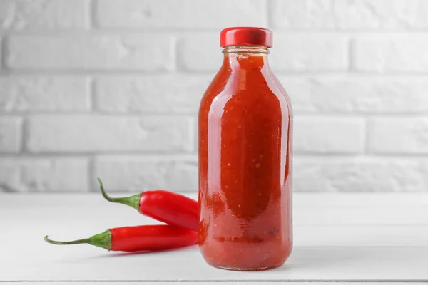 Spicy chili sauce in bottle and peppers on white wooden table