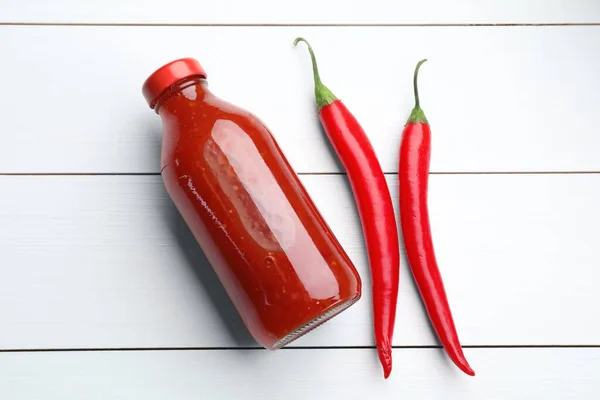 Spicy chili sauce in bottle and peppers on white wooden table, flat lay