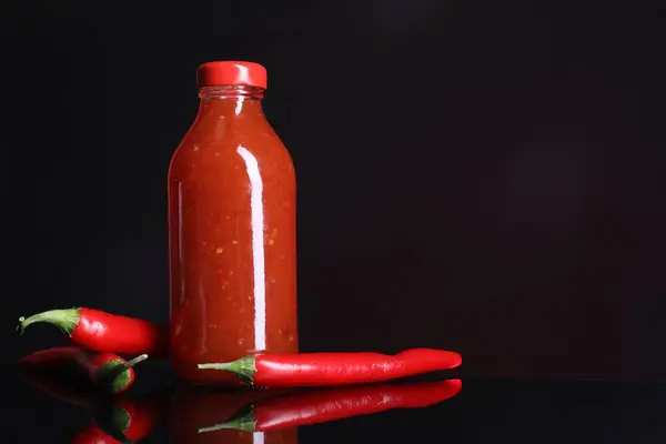 Spicy chili sauce in bottle and peppers against dark background, space for text