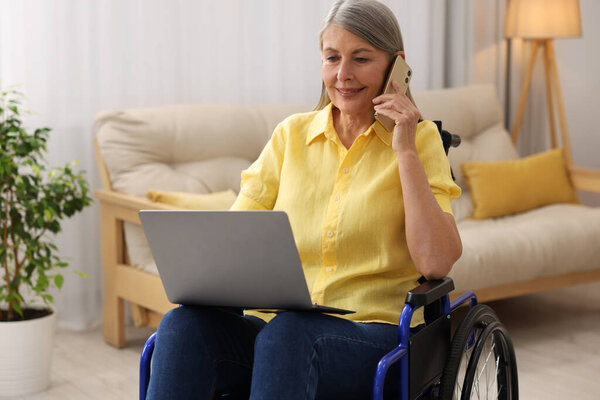 Woman in wheelchair talking on smartphone while using laptop at home