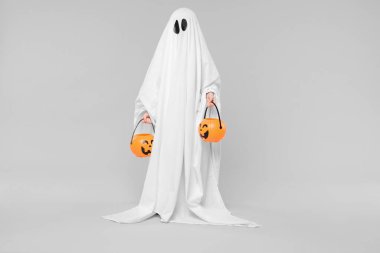 Beyaz hayalet kostümlü çocuk açık gri arka planda balkabağı kovaları tutuyor. Cadılar Bayramı