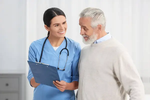 Smiling nurse with clipboard assisting elderly patient in hospital