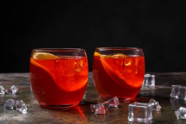 Aperol sprey kokteyli, buz küpleri ve gri desenli masada portakal dilimleri.