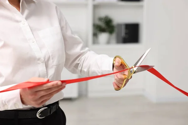 Woman cutting red ribbon with scissors indoors, closeup