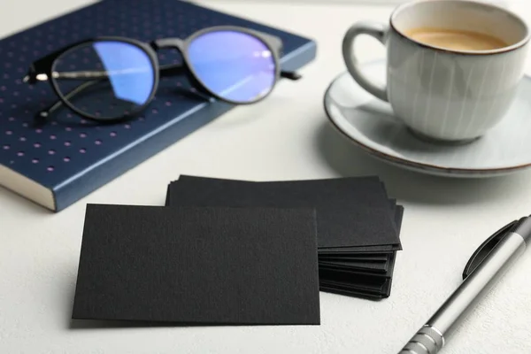 Blank black business cards, cup of coffee, glasses and stationery on white table, closeup. Mockup for design